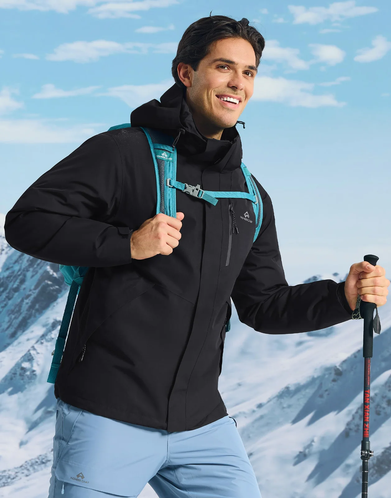 A man on a snow slope in a black hooded shell with a pale blue pack, a pole in one hand, blue sky and mountains behind him